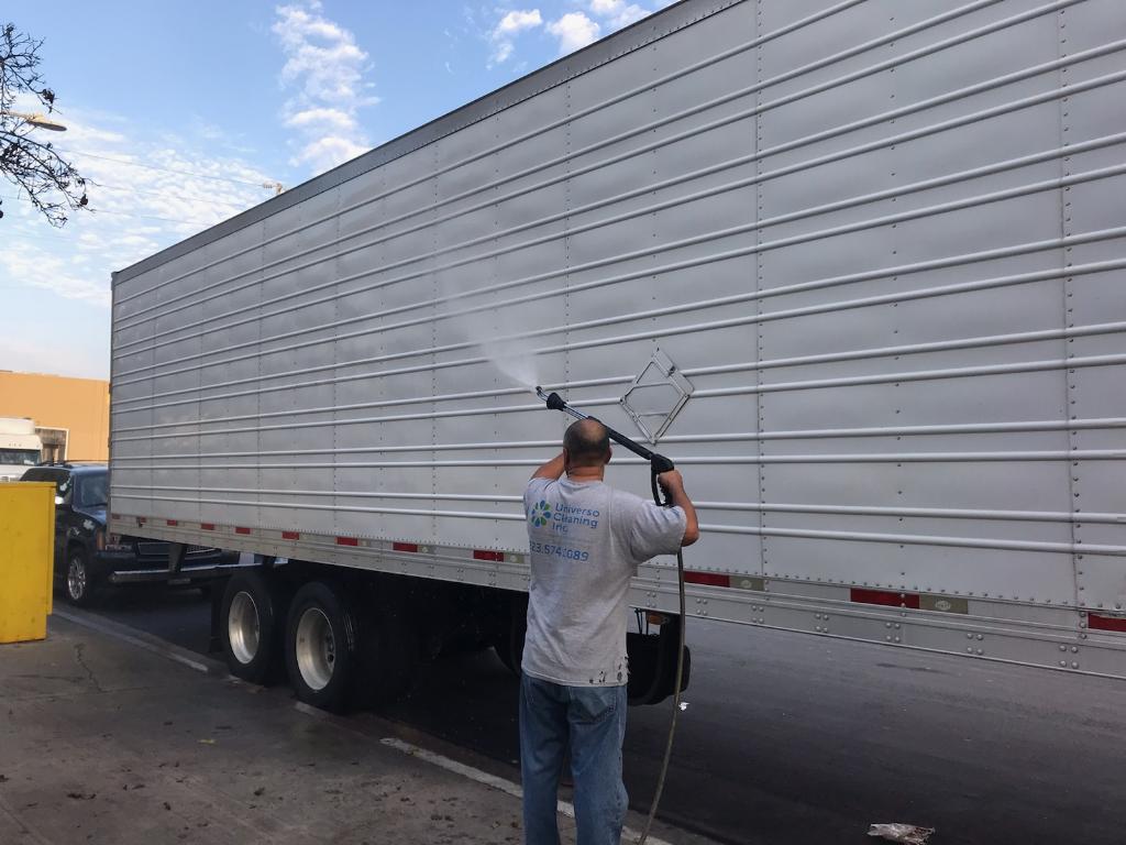 A worker sprays down a large white truck trailer with a pressure washer on a city street