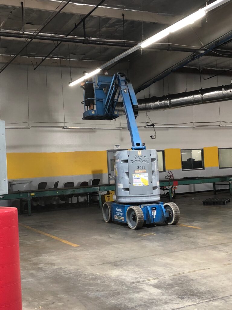 Worker on a lift platform performing maintenance on overhead lighting in an industrial space