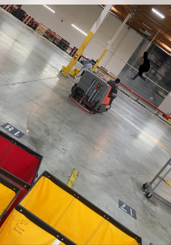 Warehouse workers operate a large floor scrubber in an industrial space