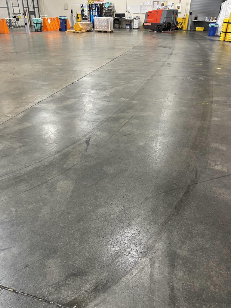 Warehouse interior showing a smooth concrete floor with visible tire marks leading towards storage areas and equipment